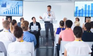 An employee meeting with a presenter at the front and charts on screens behind him.