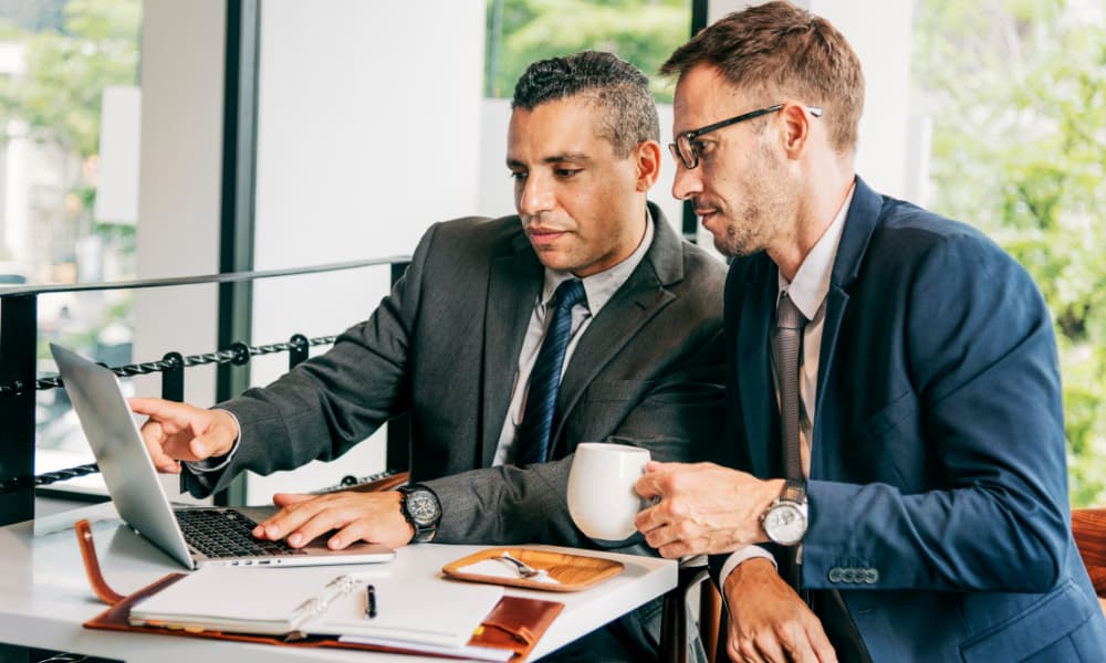 Two businessmen sit in front of a laptop while one gestures to the screen to make a point.