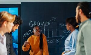 A team of people having a stand-up meeting in an office in front of a blackboard. A woman is speaking, holding a piece of chalk in her hand. The word growth and a bar chart are drawn on the blackboard.