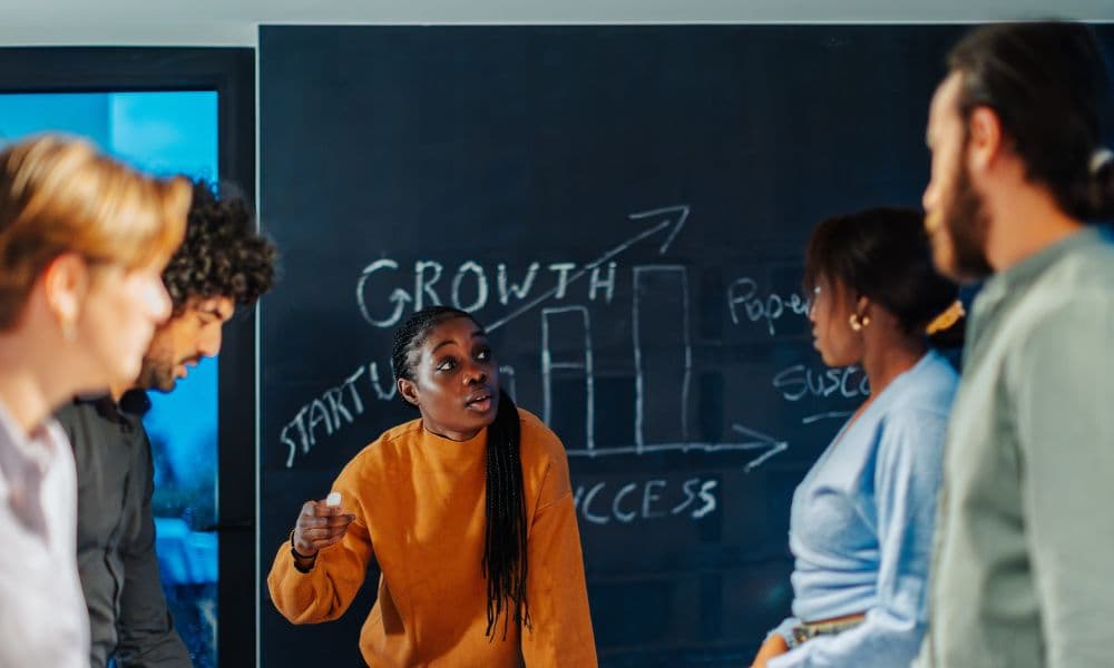A team of people having a stand-up meeting in an office in front of a blackboard. A woman is speaking, holding a piece of chalk in her hand. The word growth and a bar chart are drawn on the blackboard.