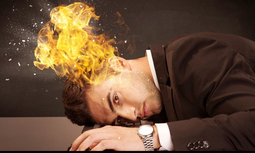 A burned-out businessman rests his head on his desk. Fire is coming from the top of his head.