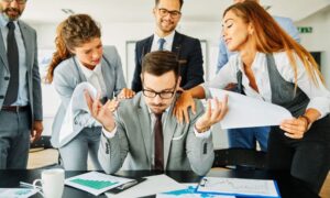 A man in a grey suit looks stressed at a desk covered in documents with charts and graphs. He is surrounded by a group of people offering documents and demanding attention.