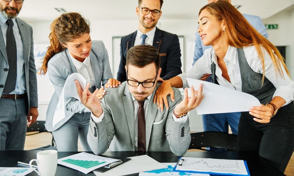 A man in a grey suit looks stressed at a desk covered in documents with charts and graphs. He is surrounded by a group of people offering documents and demanding attention.
